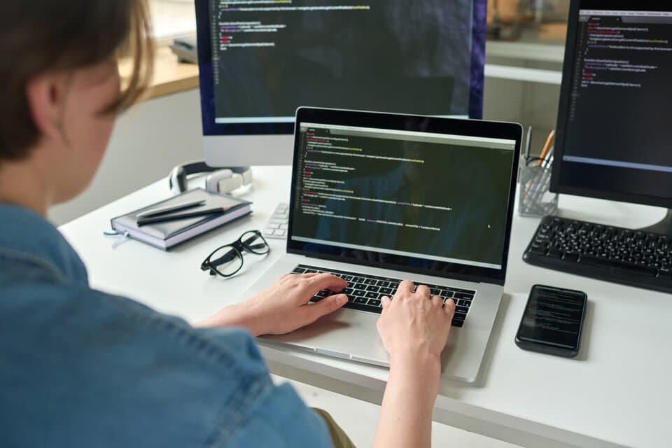 Pessoa programadora Python escrevendo código em frente ao um notebook e duas telas.
